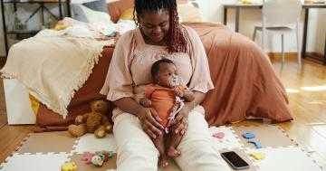 Woman on the ground leaning against a bed with a baby in her lap, representing the changes in a postpartum menstrual cycle.