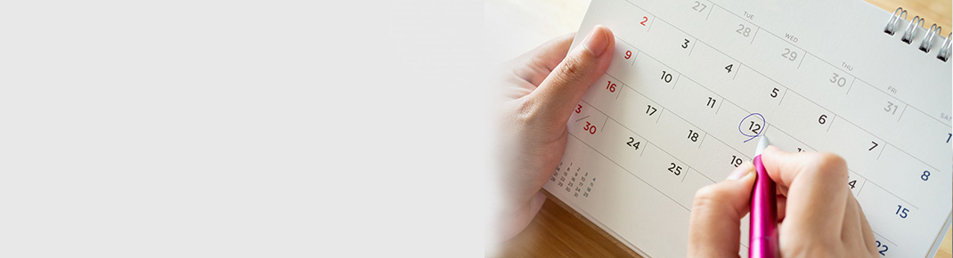 Woman in glasses wearing a dark blue shirt leans on a white bed with a smile, looking pensive. / Close up of a calendar with a woman’s hand marking a day with pen